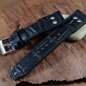 May include: Black leather watch strap with a crocodile pattern and silver buckle. The strap has stitching and multiple holes for adjusting the fit.