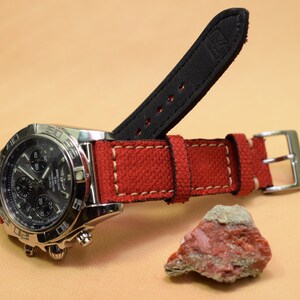 May include: A stainless steel chronograph watch with a black dial and red canvas strap. The strap has white stitching and a black leather backing. A small, reddish-brown rock sits nearby.