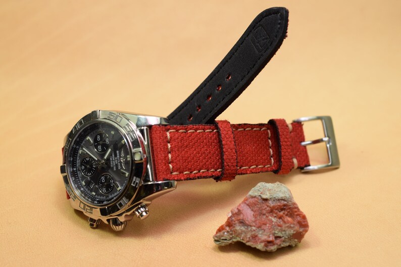 May include: A stainless steel chronograph watch with a black dial and red canvas strap. The strap has white stitching and a black leather backing. A small, reddish-brown rock sits nearby.