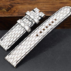 Può includere: Cinturino per orologio in due pezzi, bianco e grigio, con motivo a scaglie. Il cinturino ha cuciture nere e un lato inferiore nero. La sezione della fibbia è a sinistra e il cinturino con i fori è a destra. Lo sfondo è una superficie scura e un blocco di legno.