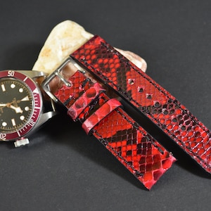 Op de afbeelding: Een polshorloge met een zwarte wijzerplaat en een rode rand, gecombineerd met een levendige rode slangenleren horlogeband. De band heeft zwarte stiksels en een zilveren gesp, wat een luxueus ontwerp laat zien.