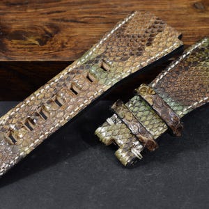 Peut inclure: Bracelet de montre en deux parties en peau de serpent. Le bracelet est principalement marron avec des accents verts et beiges, et des coutures blanches. Le bracelet comporte plusieurs trous de réglage et une boucle.