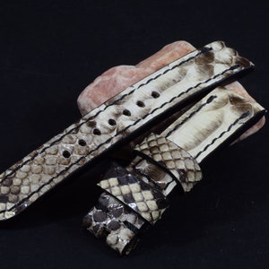 Puede incluir: Una correa de reloj de piel de serpiente blanca y gris con costuras negras. La correa está doblada por la mitad y los extremos son visibles.
