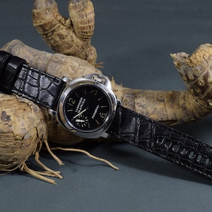 Könnte beinhalten: Ein schwarzes Lederarmband mit Krokodilprägung. Das Armband ist an einer silbernen Uhr mit schwarzem Zifferblatt und goldenen Schriftzügen befestigt, die "Luminor Marina" und "Panerai" anzeigen.