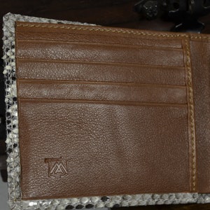May include: Open brown leather wallet with a snakeskin trim. The interior has multiple card slots and a bill compartment. The wallet has a stitched design and a small logo on the leather.