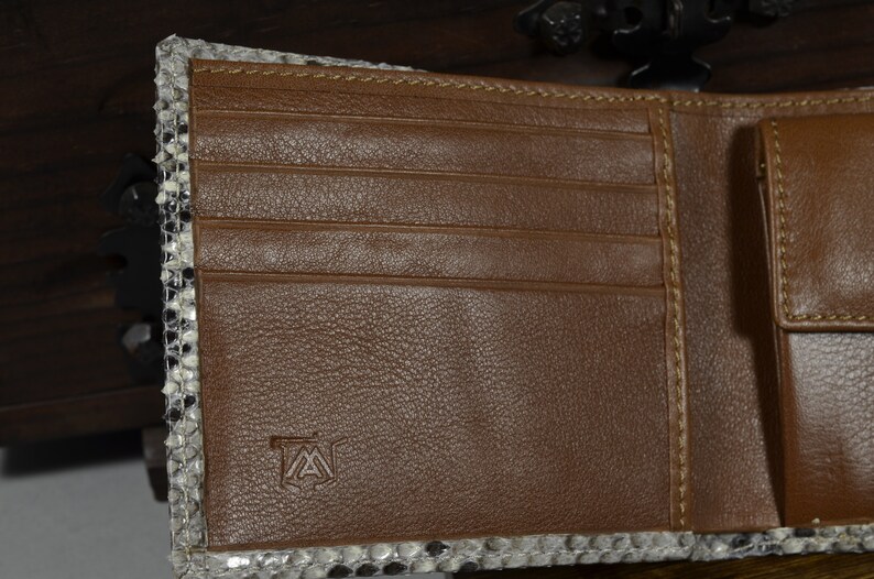 May include: Open brown leather wallet with a snakeskin trim. The interior has multiple card slots and a bill compartment. The wallet has a stitched design and a small logo on the leather.