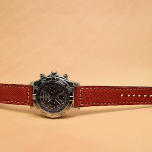 May include: A stainless steel wristwatch with a dark face and a red canvas strap. The watch features a silver buckle and white stitching along the edges of the strap. The watch is displayed on a light tan surface.
