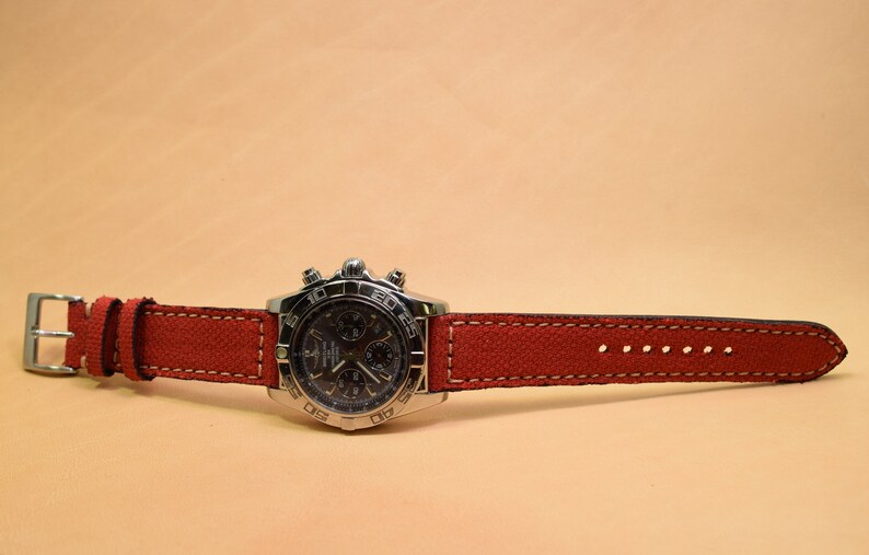 May include: A stainless steel wristwatch with a dark face and a red canvas strap. The watch features a silver buckle and white stitching along the edges of the strap. The watch is displayed on a light tan surface.
