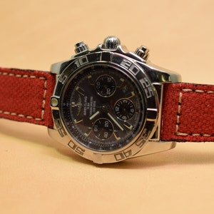 May include: A stainless steel Breitling watch with a black face and a red canvas strap. The watch features multiple subdials and a bezel with numerical markings. The red strap is stitched with white thread.