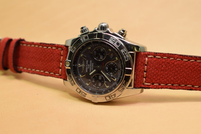 May include: A stainless steel Breitling watch with a black face and a red canvas strap. The watch features multiple subdials and a bezel with numerical markings. The red strap is stitched with white thread.