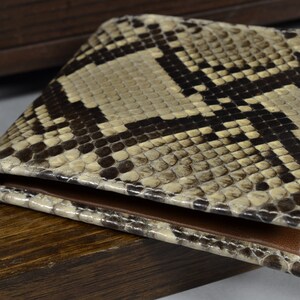 May include: A brown and beige snakeskin wallet with a textured pattern. The wallet is closed and lying on a wooden surface.