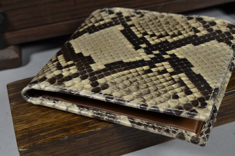 May include: A brown and beige snakeskin wallet with a textured pattern. The wallet is closed and lying on a wooden surface.