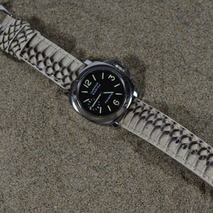 Puede incluir: Una correa de reloj de cuero blanco con un patrón de piel de serpiente blanco y negro. La correa está unida a un reloj plateado con una esfera negra y números blancos.