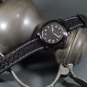 Può includere: Un orologio da polso nero con un cinturino in pelle nera e un quadrante nero con numeri romani bianchi. L'orologio è appoggiato su un oggetto in metallo argentato.