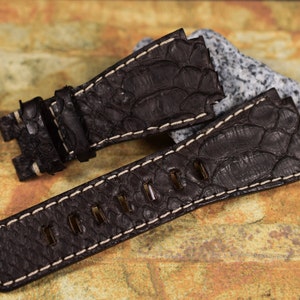 May include: A dark brown leather watch strap with white stitching. The strap is made of a textured material that resembles alligator skin.