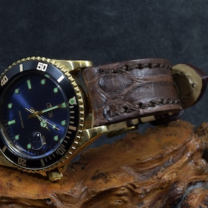 Può includere: Un orologio dorato con quadrante blu e ghiera nera. L'orologio ha un cinturino in pelle marrone con motivo coccodrillo.