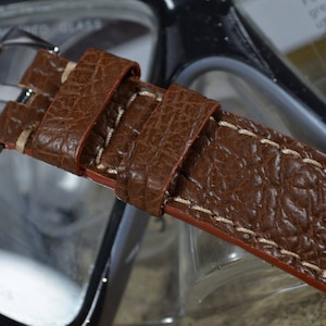 May include: A brown leather watch strap with white stitching. The strap has a textured surface and a silver buckle.