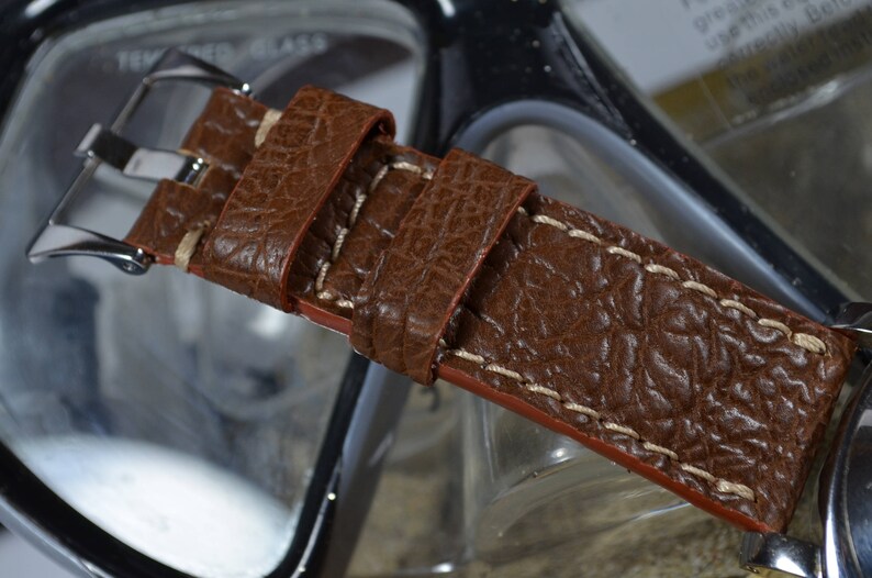 May include: A brown leather watch strap with white stitching. The strap has a textured surface and a silver buckle.