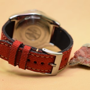 May include: Close-up of a wristwatch with a red canvas strap and a silver buckle. The watch has a black leather backing and a silver case. A small, reddish-brown rock sits to the right of the watch.