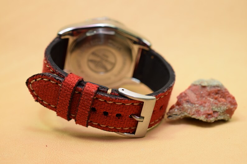 May include: Close-up of a wristwatch with a red canvas strap and a silver buckle. The watch has a black leather backing and a silver case. A small, reddish-brown rock sits to the right of the watch.