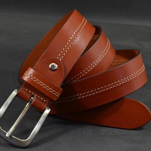 May include: A brown leather belt with a silver buckle. The belt is stitched with white thread and has a classic design. The belt is rolled up, showing the details of the leather and stitching.