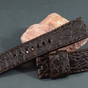 Puede incluir: Una correa de reloj de piel de cocodrilo marrón con borde cosido y hebilla.