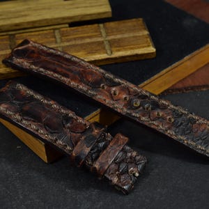 May include: Two brown leather watch straps with a reptile skin pattern. The straps are dark brown with lighter brown stitching and a buckle. The straps are lying on a wooden surface.