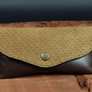 May include: A brown leather glasses case with a tan snakeskin pattern on the front flap. The case has a brass button closure and a small logo on the front.