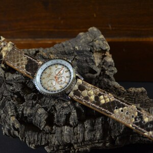 Può includere: Un orologio cronografo argentato con quadrante bianco e lancette nere. L'orologio è su un cinturino in pelle con motivo a pelle di serpente marrone e nero.