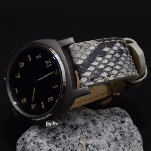 Peut inclure: Une montre-bracelet à cadran noir avec des chiffres romains pour les heures et un bracelet en cuir à motif de peau de serpent blanc.