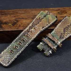 Könnte beinhalten: Zweiteiliges Uhrenarmband-Set. Die Armbänder weisen ein Schlangenmuster in Grün-, Braun- und Beigetönen auf. Die Kanten sind mit weißem Faden genäht. Die Uhrenarmbänder werden auf einer dunklen Holzoberfläche präsentiert.