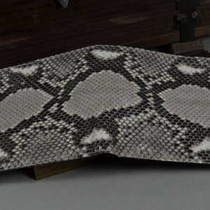 May include: A bi-fold wallet with a snakeskin pattern in shades of grey, black, and white. The wallet is open, revealing the interior. The texture of the snakeskin is visible. The wallet is likely made of genuine leather.