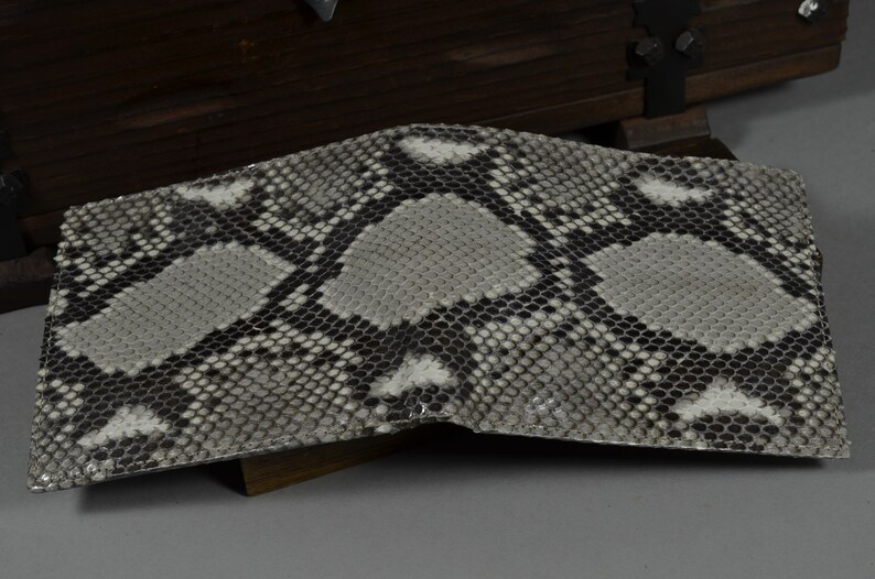 May include: A bi-fold wallet with a snakeskin pattern in shades of grey, black, and white. The wallet is open, revealing the interior. The texture of the snakeskin is visible. The wallet is likely made of genuine leather.