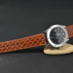 Könnte beinhalten: Ein braunes Lederarmband mit Krokodilmuster. Das Armband ist an einer silbernen Uhr mit schwarzem Zifferblatt und silbernen Zeigern befestigt.