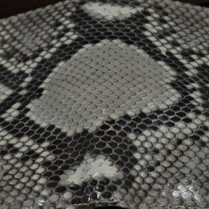 May include: Close-up of a textured surface with a snakeskin pattern. The design features a repeating pattern of scales in shades of grey, black, and white. The scales are small and tightly packed, creating a detailed and visually interesting texture.