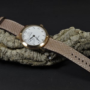 Peut inclure: Une montre en or rose avec un cadran blanc et des aiguilles noires. La montre a un bracelet en cuir brun avec un motif texturé. La montre est posée sur un morceau d'écorce.