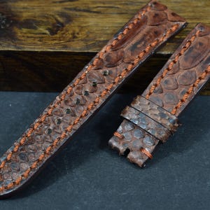 May include: Two brown watch straps with a textured, reptile-like pattern. The straps are stitched with orange thread. One strap has a buckle and the other has a long strap.