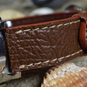 May include: A brown leather watch strap with white stitching. The strap is made of textured leather and has a smooth, red underside.