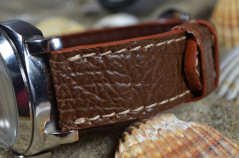 May include: A brown leather watch strap with white stitching. The strap is made of textured leather and has a smooth, red underside.
