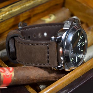 May include: A wristwatch with a dark brown suede leather strap and a polished silver-tone metal case. The watch features a black dial with luminous hands and markers, including the text Luminor Marina printed on the face of the watch.