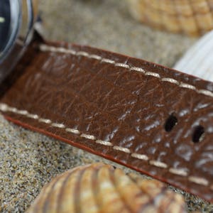 May include: Close-up of a brown leather watch strap with white stitching. The strap has a textured surface and multiple holes for adjusting the fit.