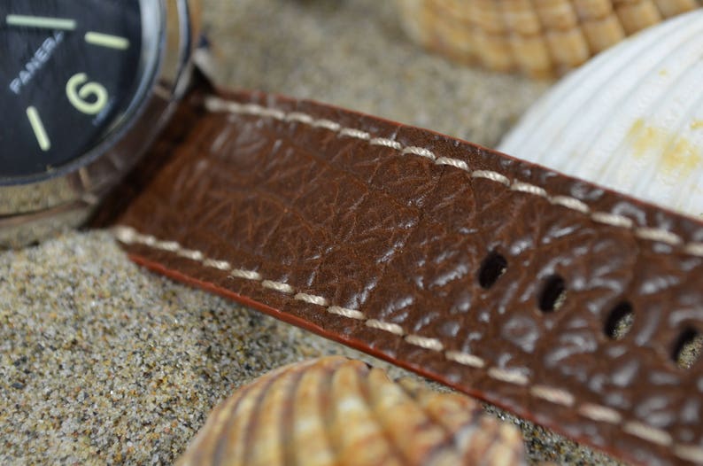 May include: Close-up of a brown leather watch strap with white stitching. The strap has a textured surface and multiple holes for adjusting the fit.