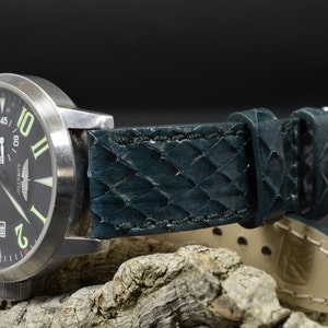 Peut inclure: Une montre noire avec un bracelet en cuir vert. Le cadran de la montre a des chiffres et des aiguilles blancs, et le bracelet a un motif texturé.