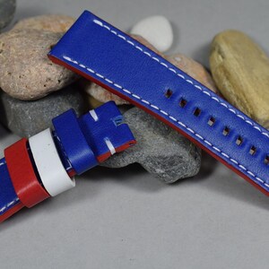 Op de afbeelding: Een blauw, rood en wit leren horlogebandje met witte stiksels. Het bandje is dubbelgevouwen, waardoor de onderkant zichtbaar is.