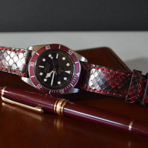 Może przedstawiać: Czarno-biały zegarek z czerwoną ramką i paskiem ze skóry węża w kolorze czerwonym i czarnym. Zegarek leży na brązowej skórzanej powierzchni obok czerwonego długopisu.