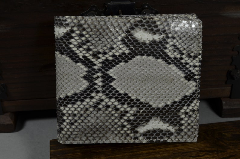 May include: A bi-fold wallet with a snakeskin pattern in shades of grey, black, and white. The wallet is made of a textured material and appears to be new. The wallet is displayed on a wooden surface.