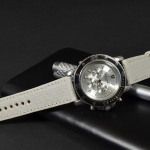 Puede incluir: Una correa de reloj de cuero blanco con un reloj cronógrafo plateado con un bisel negro. El reloj tiene una esfera blanca con marcas negras y una aguja de segundos plateada.