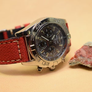 May include: A silver wristwatch with a dark face and multiple dials, paired with a red canvas strap. The watch is next to a small, reddish-brown rock. The watch strap has white stitching.