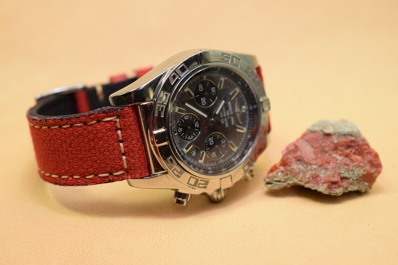 May include: A silver wristwatch with a dark face and multiple dials, paired with a red canvas strap. The watch is next to a small, reddish-brown rock. The watch strap has white stitching.