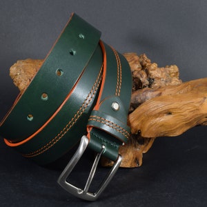 Könnte beinhalten: Ein dunkelgrüner Ledergürtel mit orangefarbenen Nähten und einer silbernen Schnalle. Der Gürtel ist aufgerollt und auf einem Stück Treibholz ausgestellt.